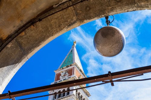 venezianischer Glockenturm der Markusbasilika in Venedig gegen den blauen Himmel aus einer ungewöhnlichen Perspektive, die Spitze des Turms mit einem in der Sonne leuchtenden Engel, umgeben von einem Steinbogen, italienische Wahrzeichen